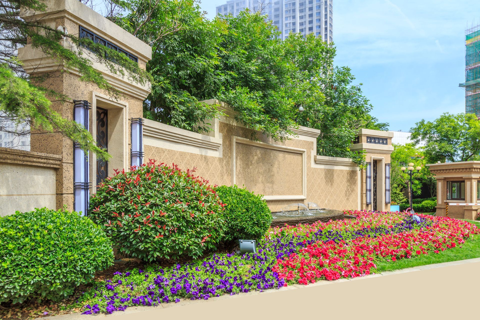 Landscaped stone wall with colorful flowers and greenery along a city sidewalk under blue sky