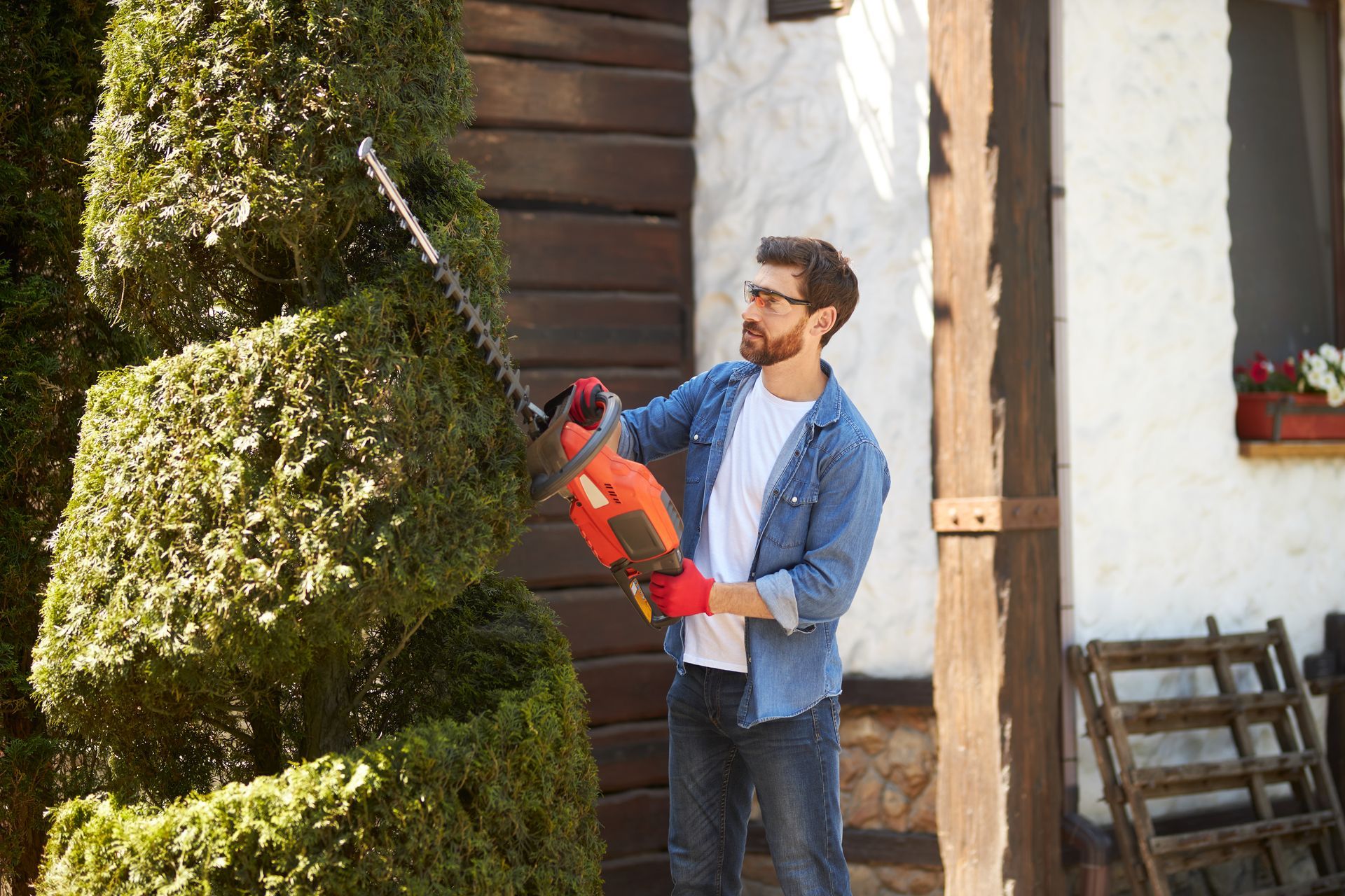 Man in denim shirt and red gloves trims a green topiary with an orange hedge trimmer outside a house.