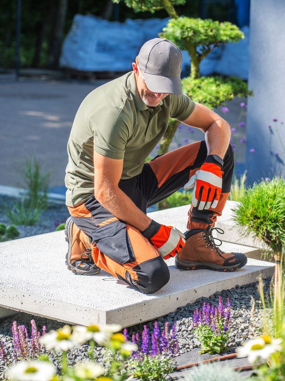 A man is kneeling down in a garden wearing gloves and boots.