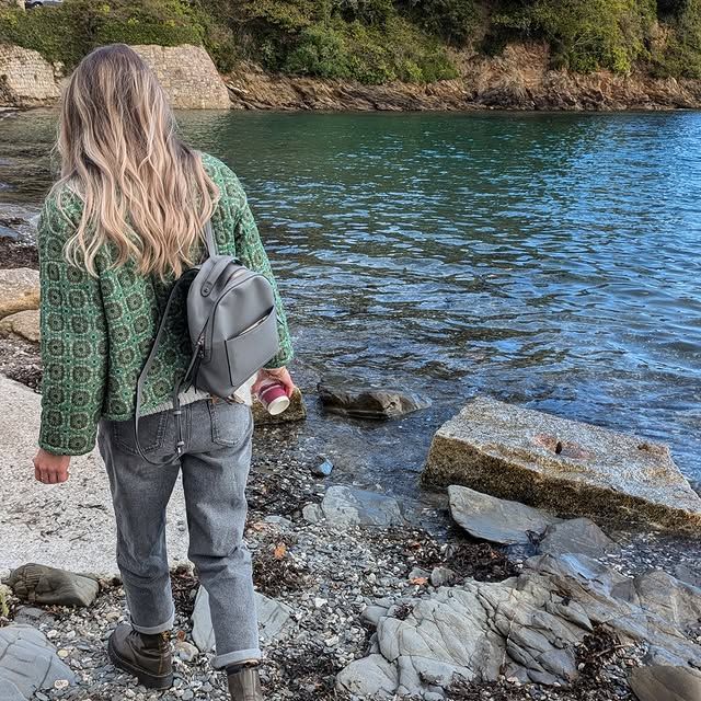 Woman walking on rocky beach, wearing green jacket and backpack, near water.