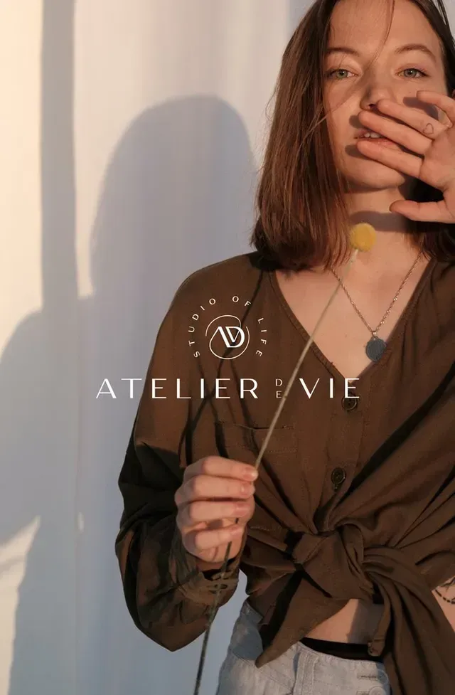 Woman holding flower, wearing brown shirt, partially covering face. Atelier de Vie logo.