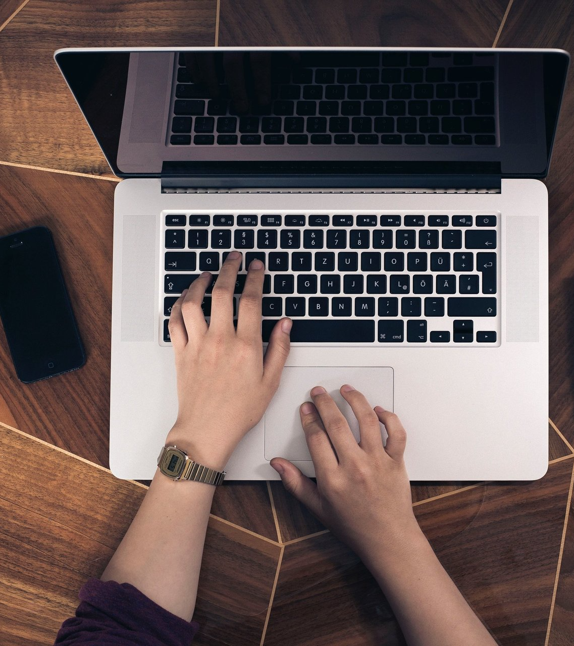 Hands typing on a laptop with a watch, phone, and dark wood surface visible.
