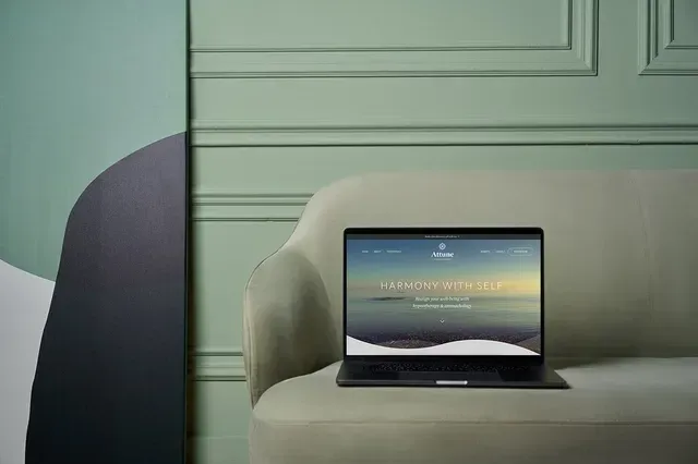 Laptop showing a website on a light-colored sofa displaying a website. Background includes light green paneling and abstract art.