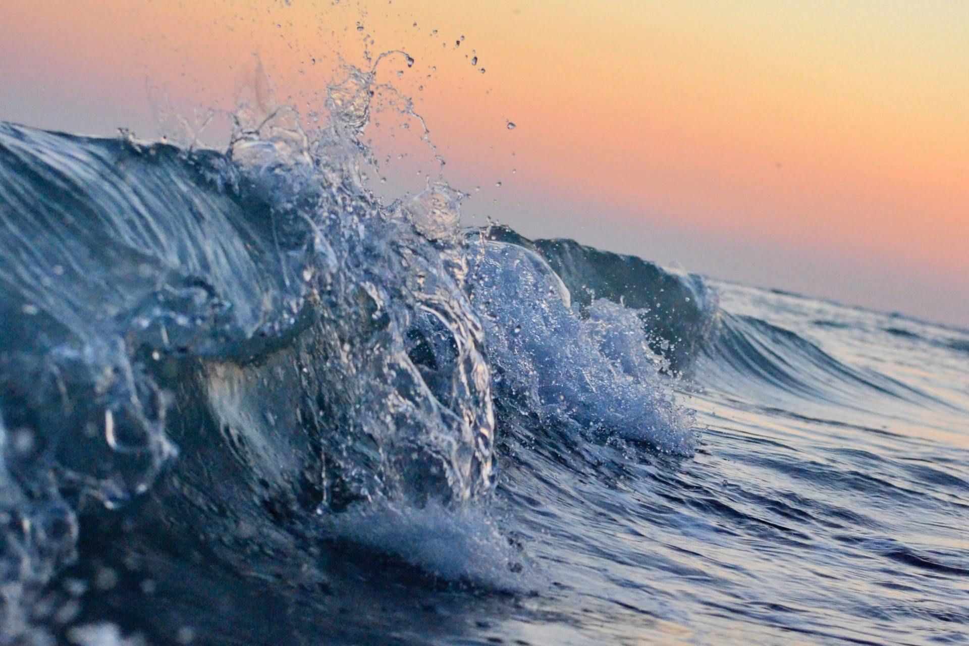 Ocean wave cresting with water splashing; sunset in the background with orange and pink hues.