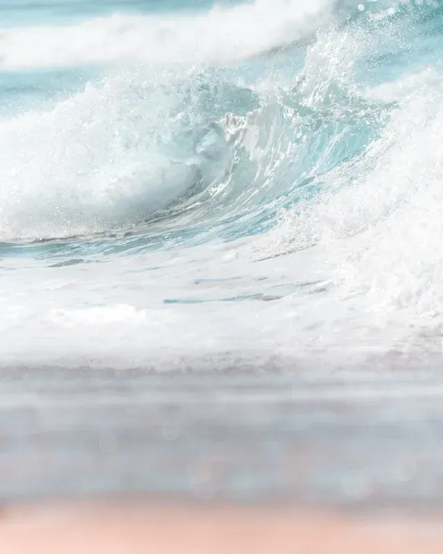 Crashing turquoise ocean wave, white foam, blurry foreground of sandy beach.