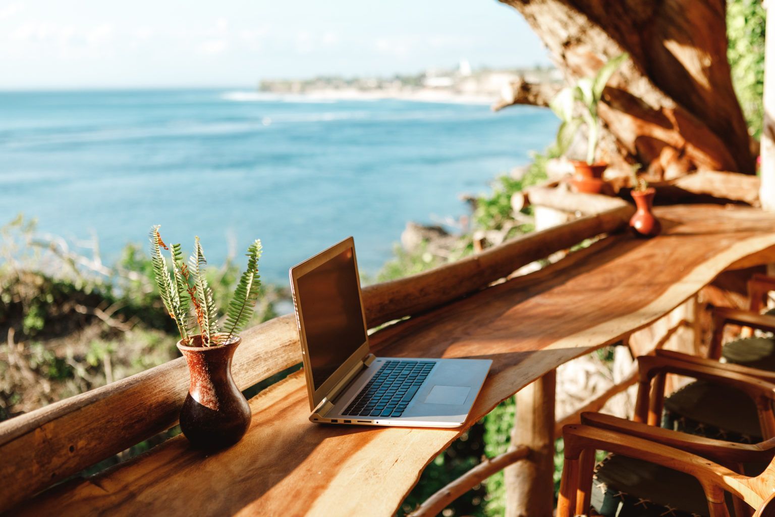 Woman in a hat works on a laptop at a wooden table overlooking the ocean.