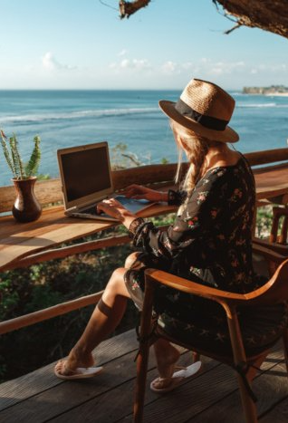 Woman in a hat works on a laptop at a wooden table overlooking the ocean.