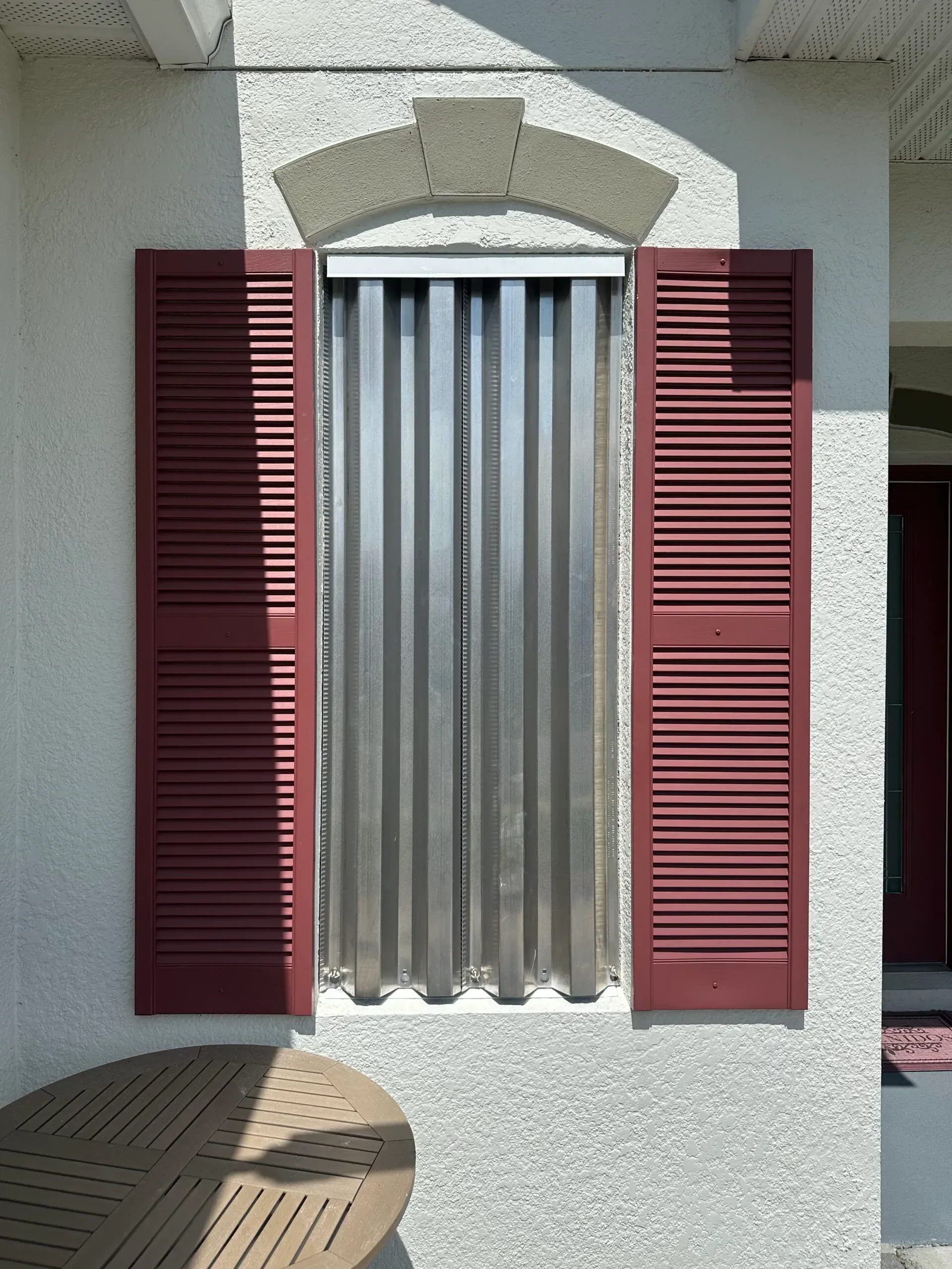 Window with accordion-style hurricane shutters closed, flanked by red shutters. Gray exterior.