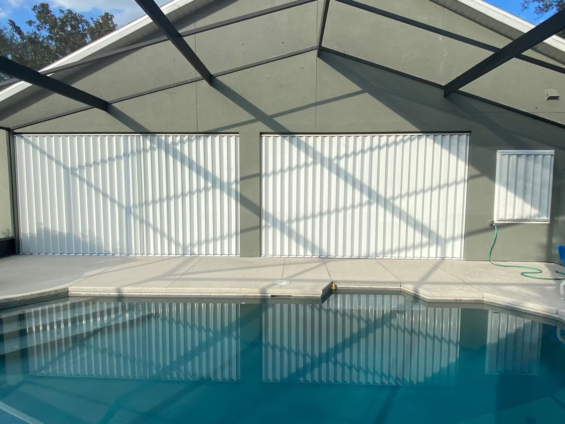 Pool area with white accordion hurricane shutters closed on a building with a dark roof frame.