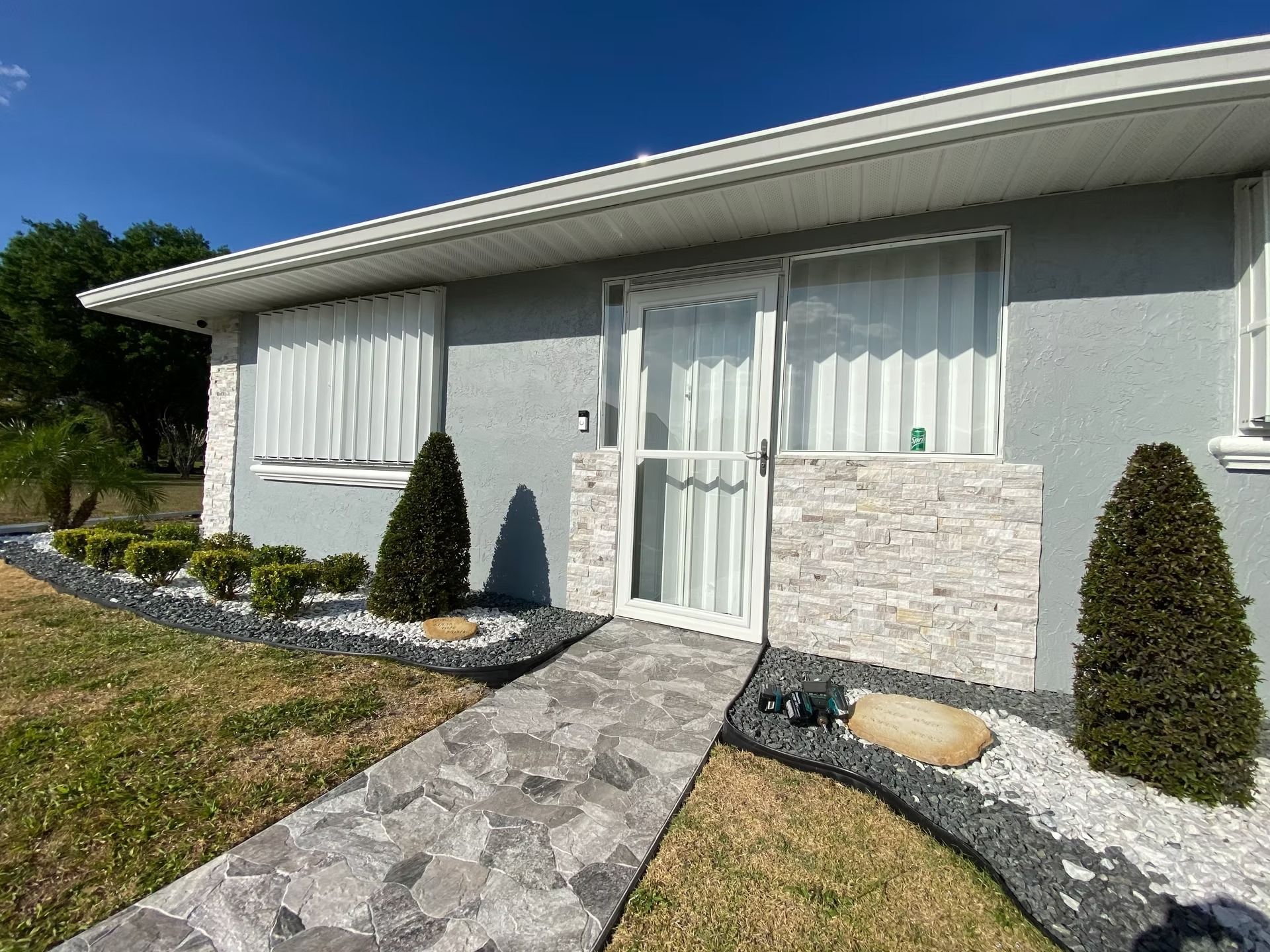 Gray house with stone walkway and facade, lush landscaping under blue sky.