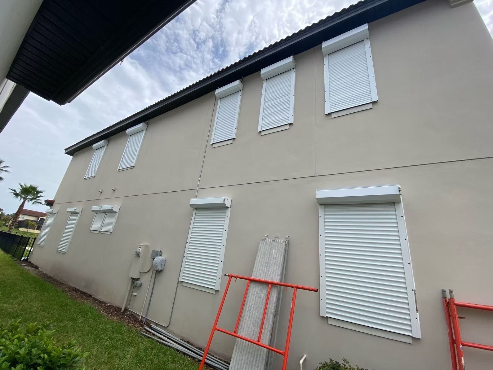Tan two-story house with white hurricane shutters, green grass, and a partly cloudy sky.