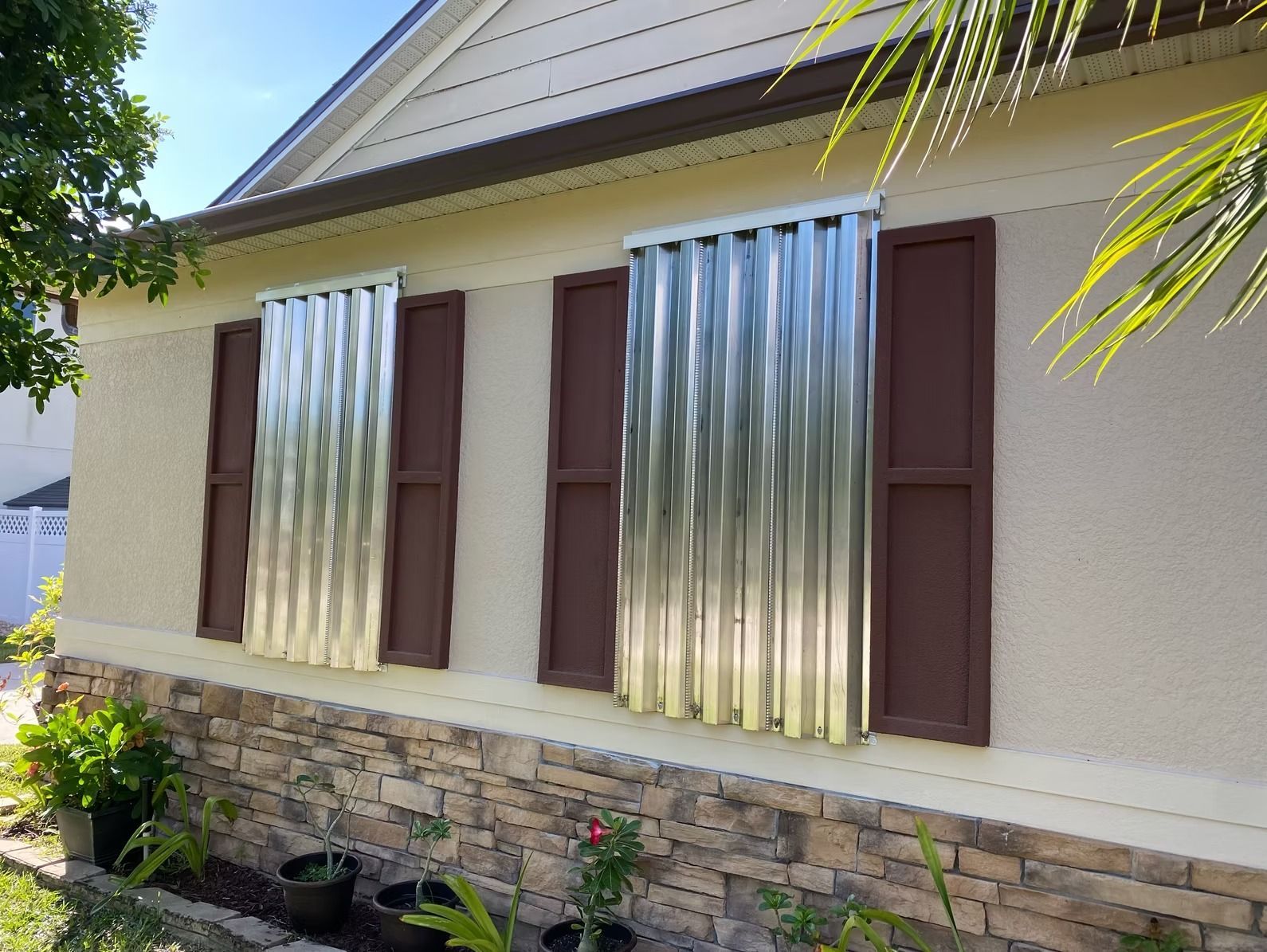 House with corrugated metal hurricane shutters over windows; brown shutters are also present.