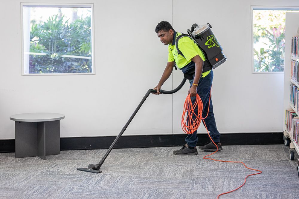 A person wearing blue gloves sprays and wipes a white office desk, cleaning with a yellow cloth.