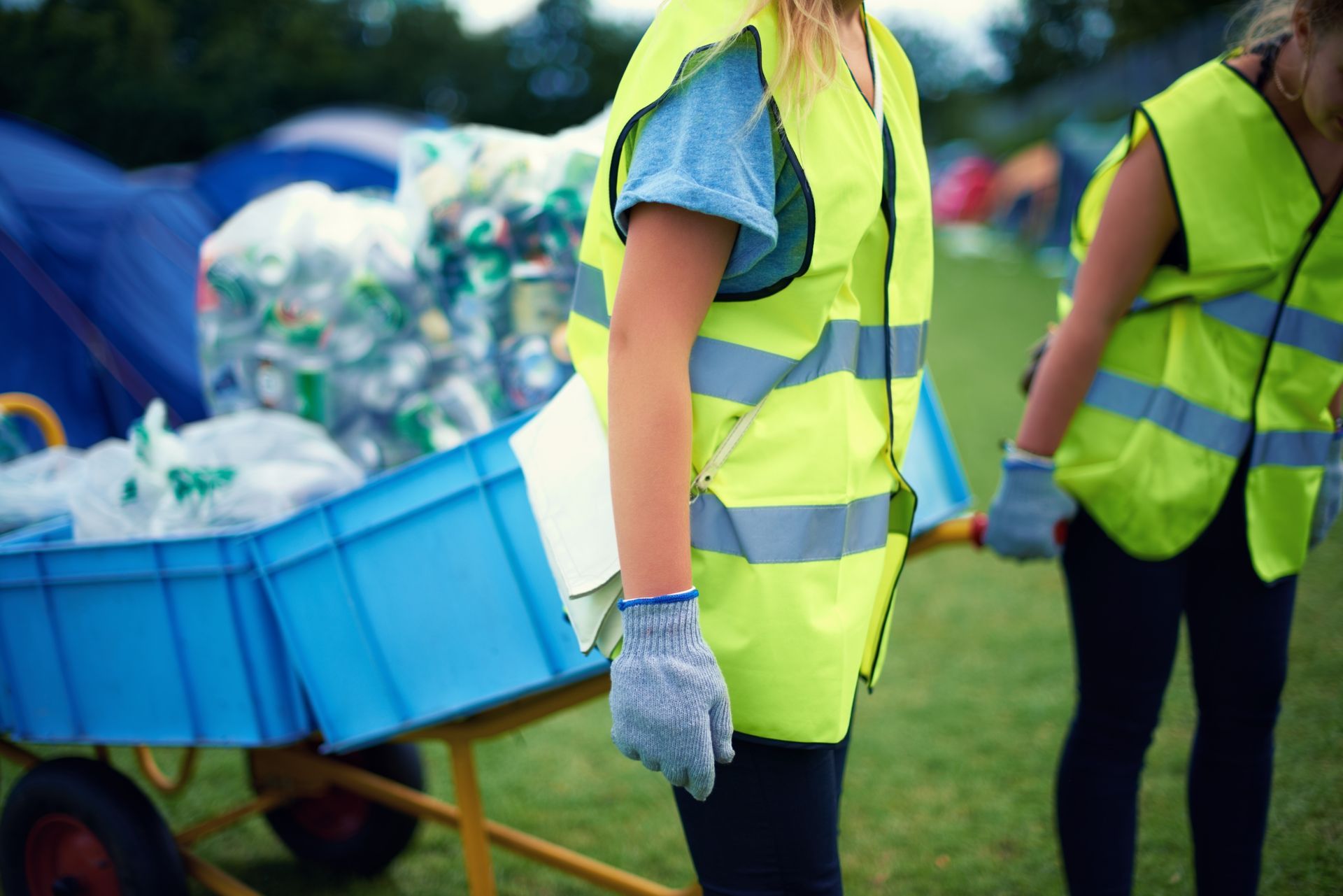 Two people in reflective vests pushing a cart of recycling bins on a grassy field.