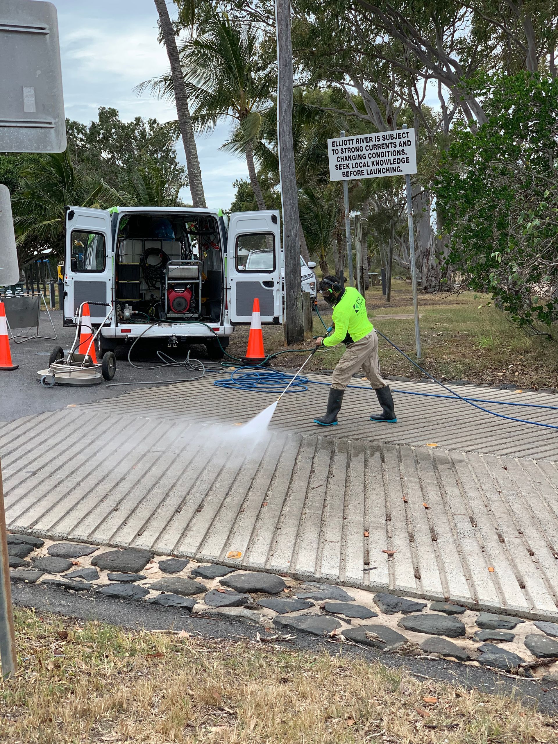 A worker in green overalls sprays a wet sidewalk. The setting is outdoors, with a small tree and concrete barriers visible.
