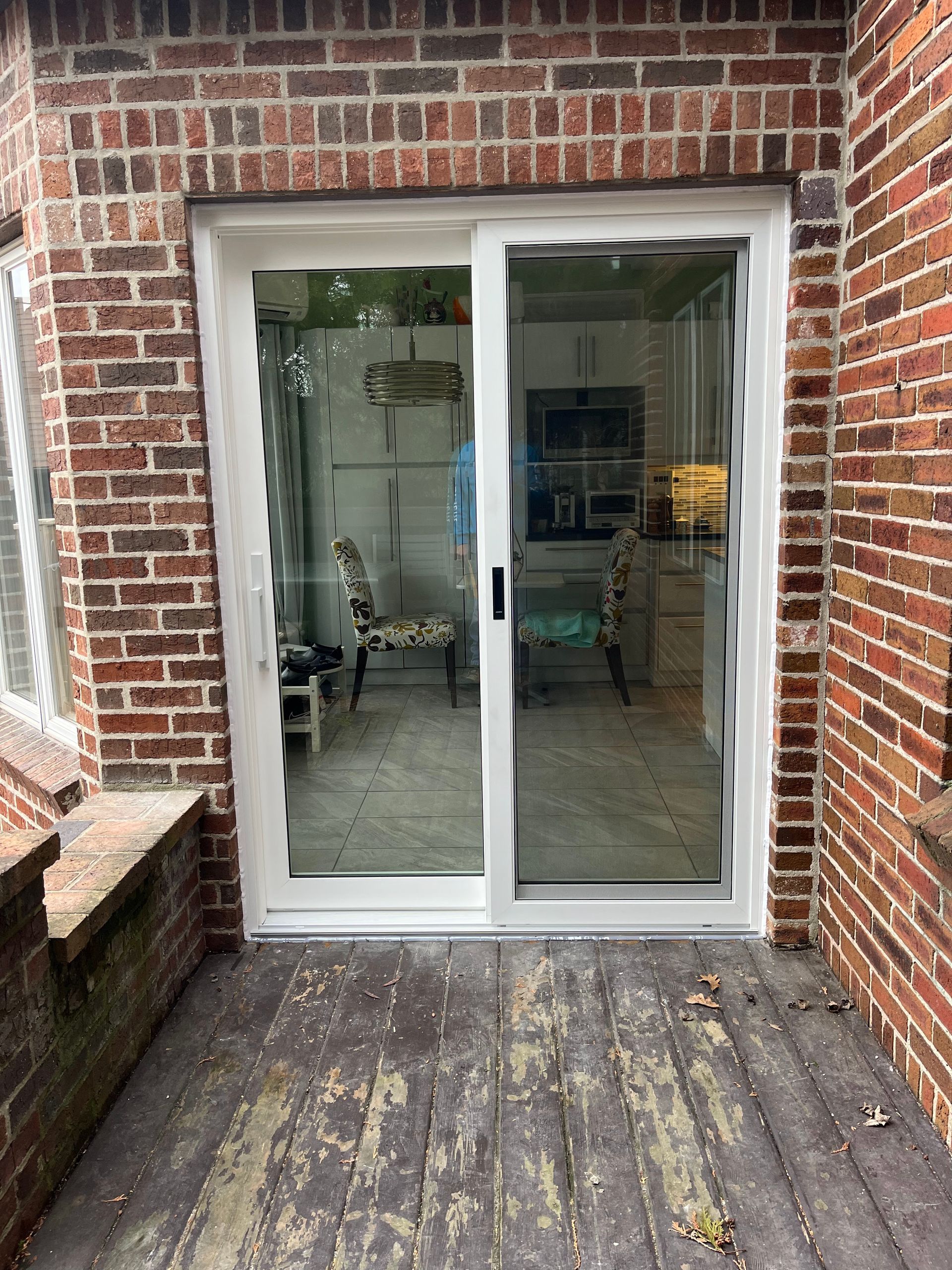 A white sliding glass door is between a brick wall and a wooden deck.