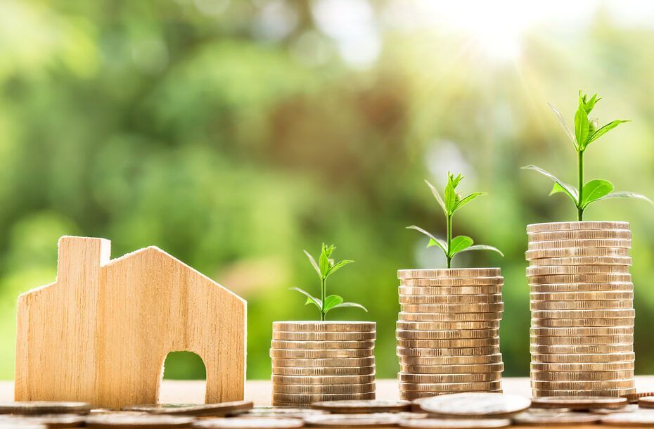 A wooden house is sitting next to stacks of coins with plants growing out of them.