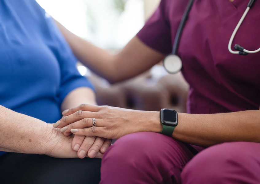 Nurse in burgundy scrubs comforts an elderly woman, holding her hand.