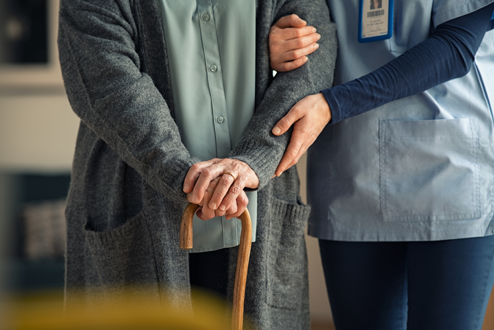 A senior woman with a cane is assisted by a caregiver; indoors.