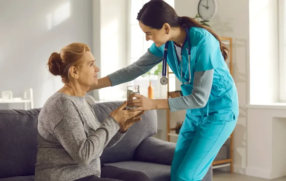 Nurse in blue scrubs offers a glass of water to an elderly woman on a couch.