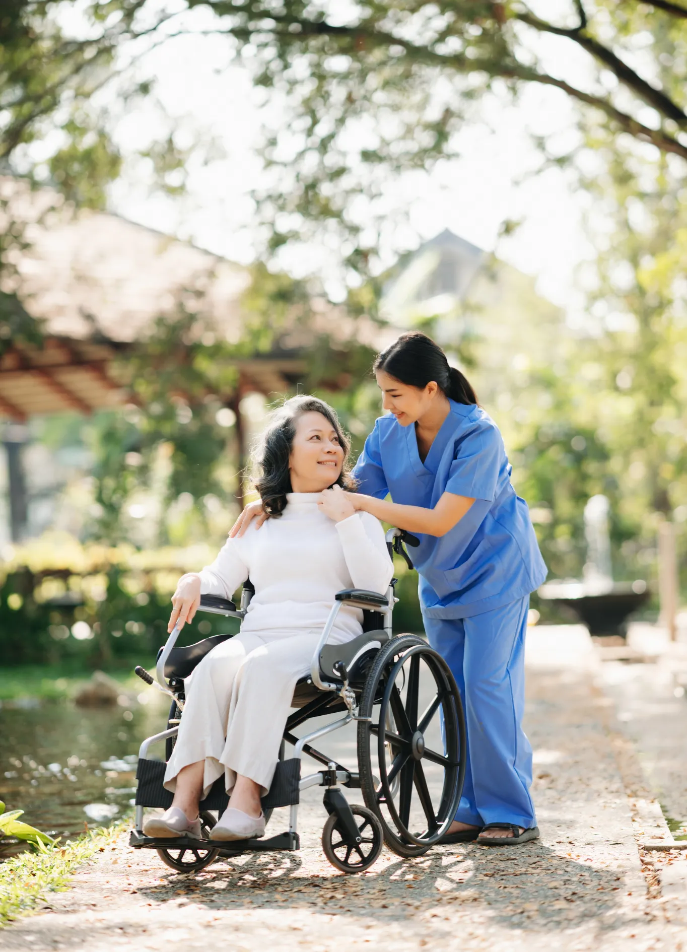 Woman in wheelchair with caregiver outside on a sunny day.