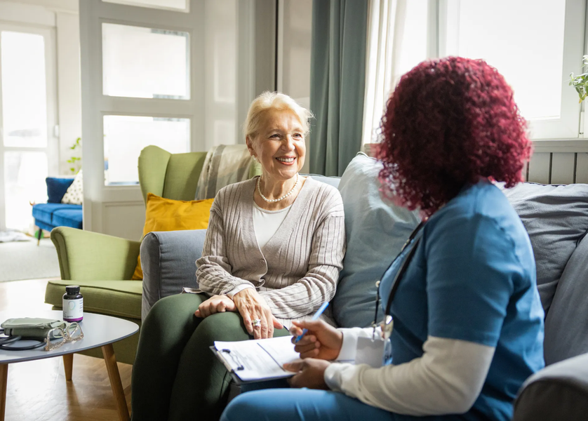 Older woman consults with healthcare worker on a sofa, discussing paperwork in a home setting.