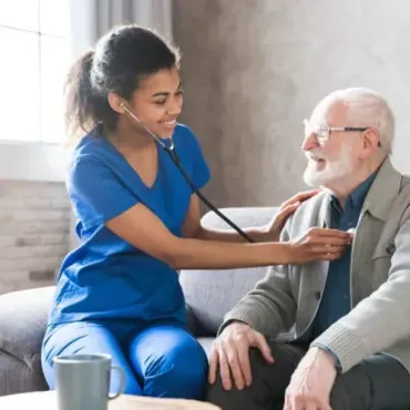 Nurse in blue scrubs listens to older man's heart with a stethoscope in a living room.