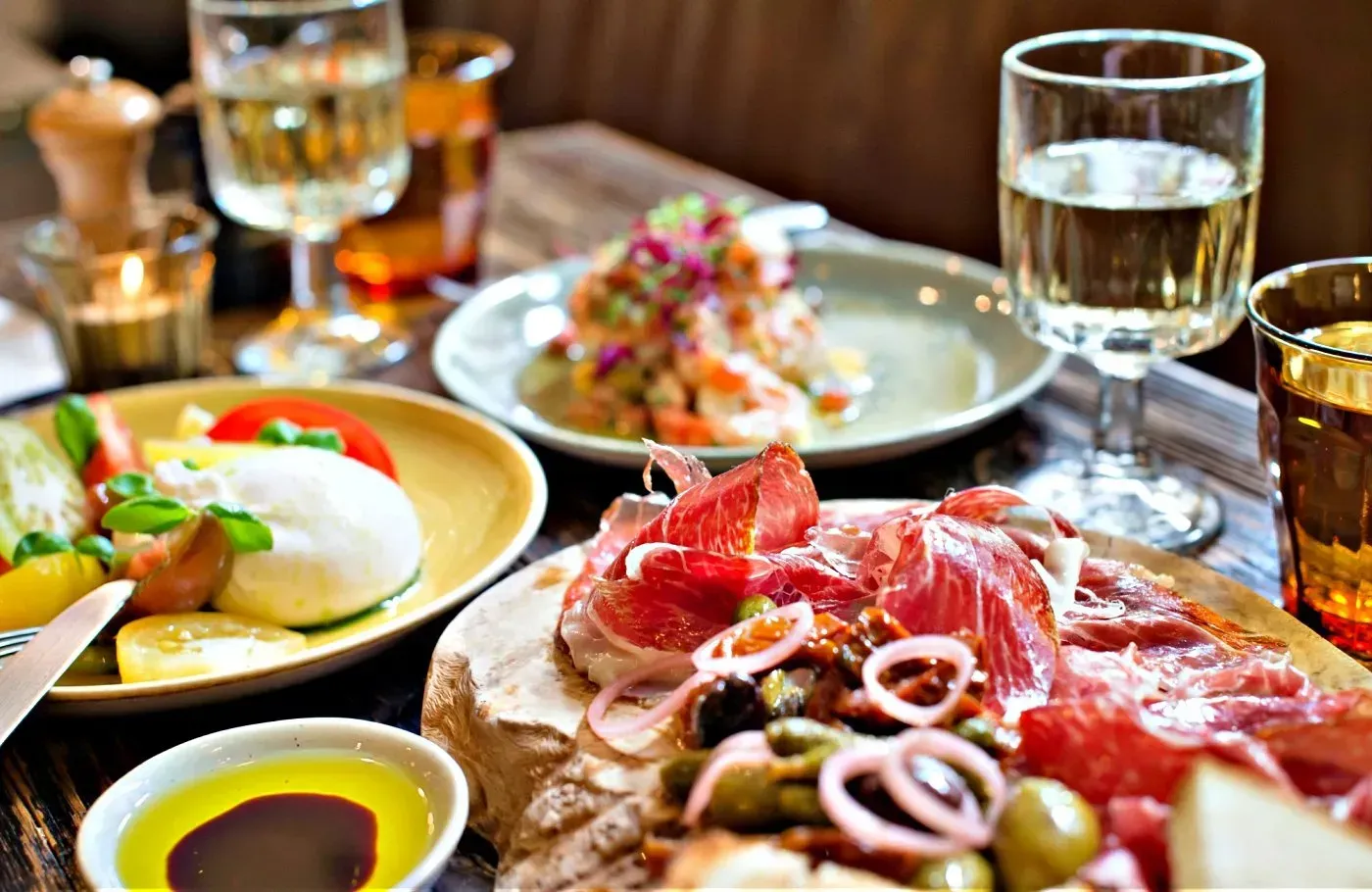 Platter of cured meats and olives, with salads and wine glasses on a table.