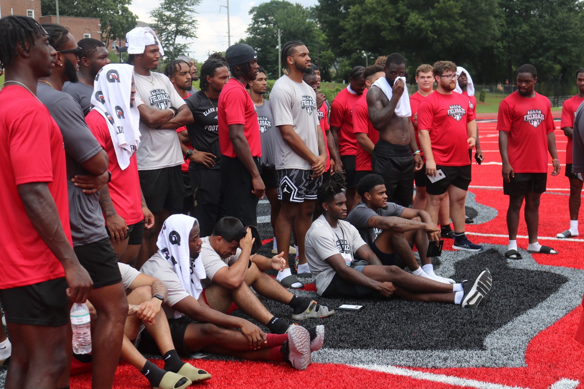 Athletes in red and grey shirts stand and sit on a red track. Some hold towels, likely a team huddle.