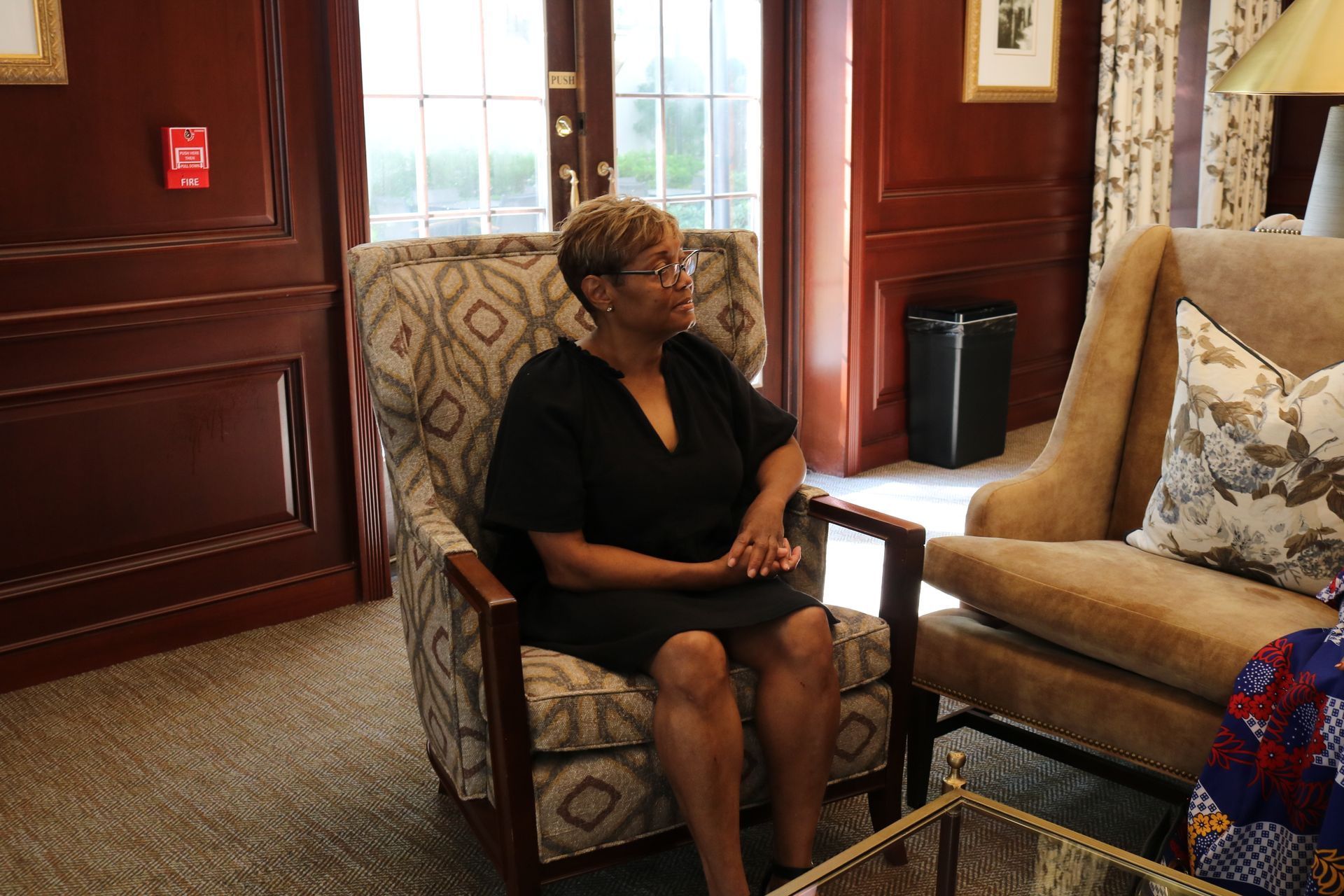 Woman in a black dress sits in a patterned armchair in a room with wood paneling and natural light.