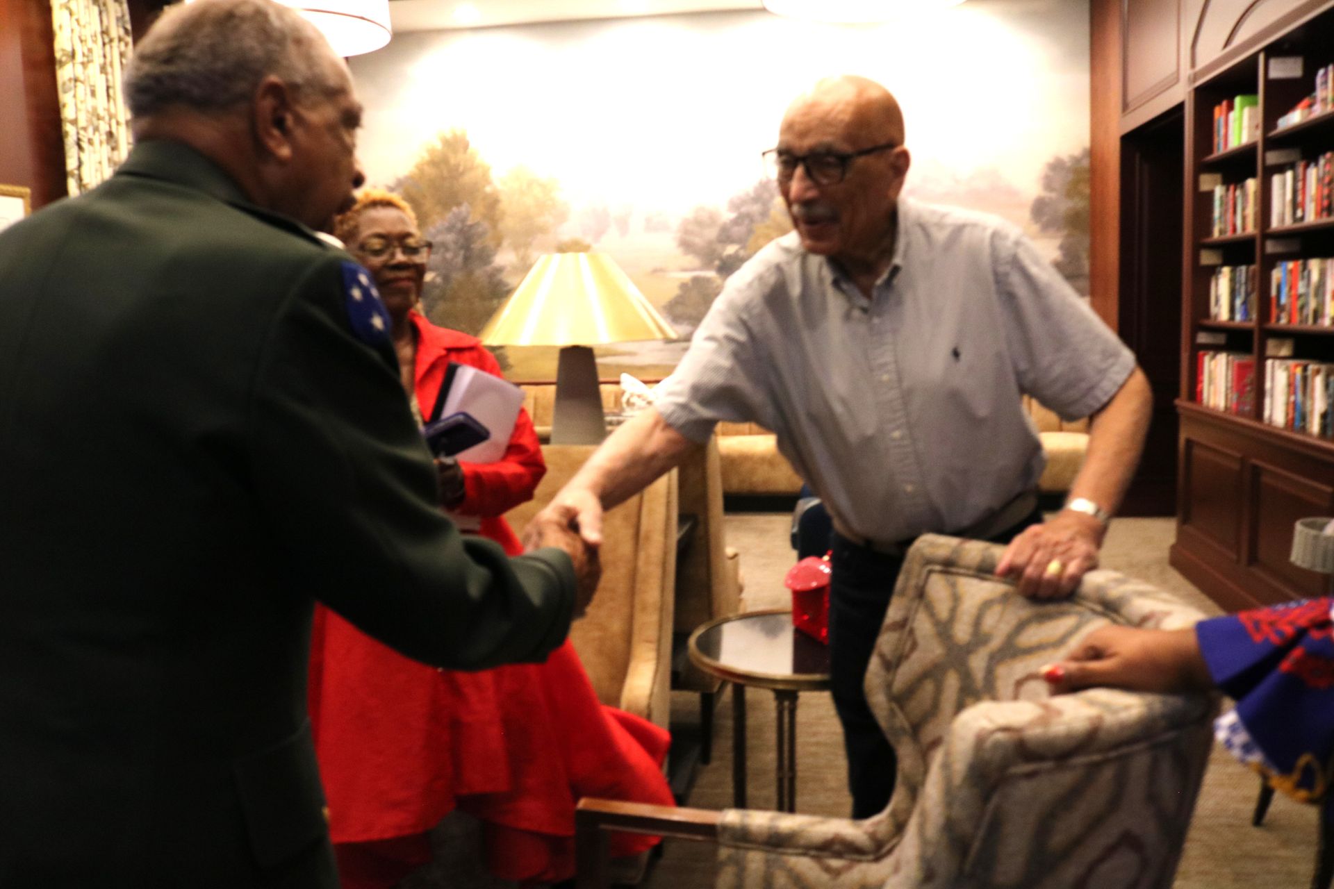 Man in olive jacket shakes hands with man in light blue shirt, near seating and bookshelves.