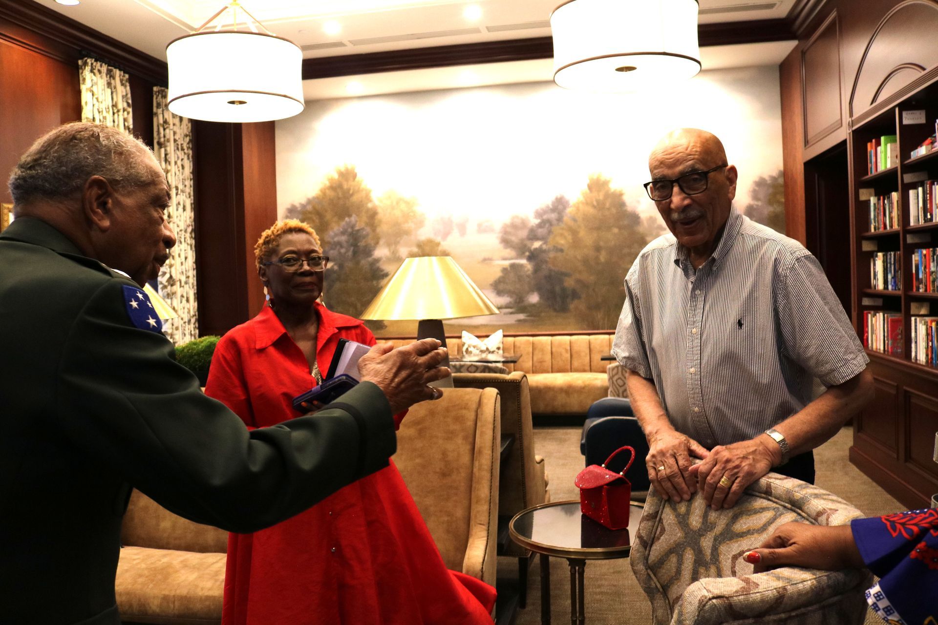 Three people inside a room with furniture, one taking a photo with their phone. Background has a mural and bookshelves.