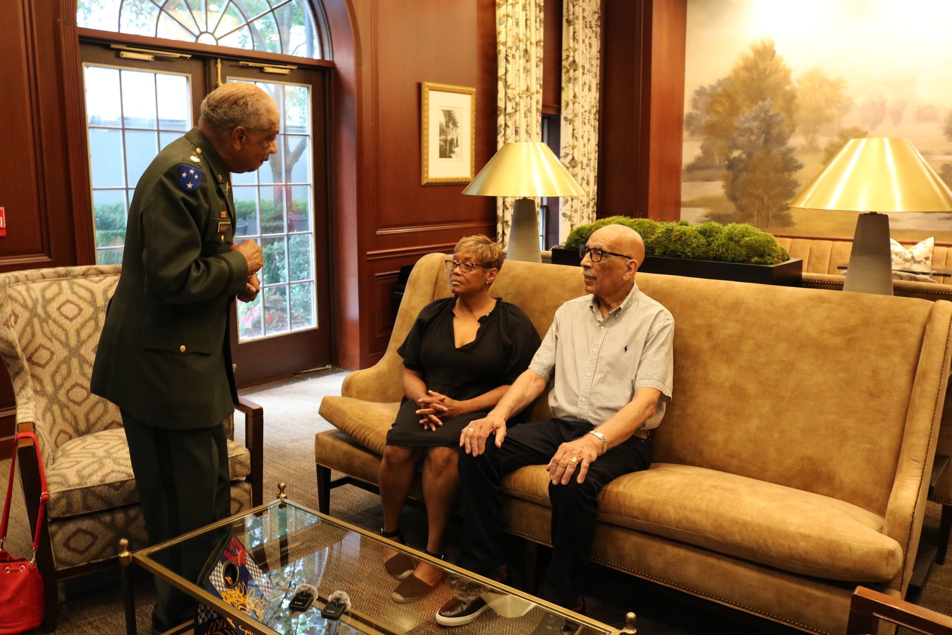 Man in uniform talking to a couple on a sofa in a lobby. Neutral colors, indoor setting.
