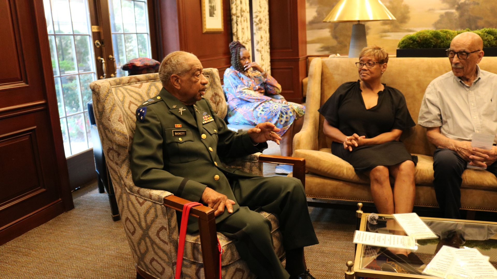 Man in military uniform seated, speaking to three others in a room; a lamp and side table are visible.