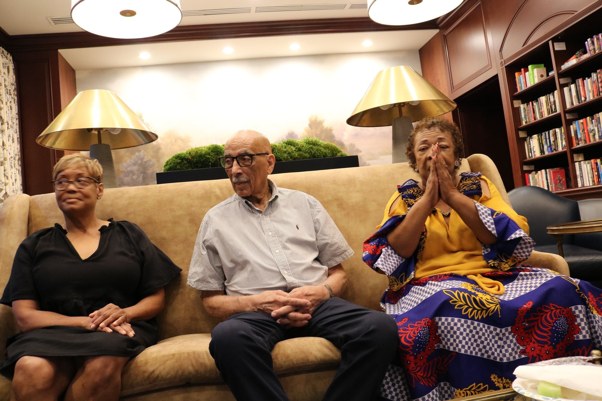 Three people seated on a couch indoors; two women, one man. The woman on the right has hands clasped.