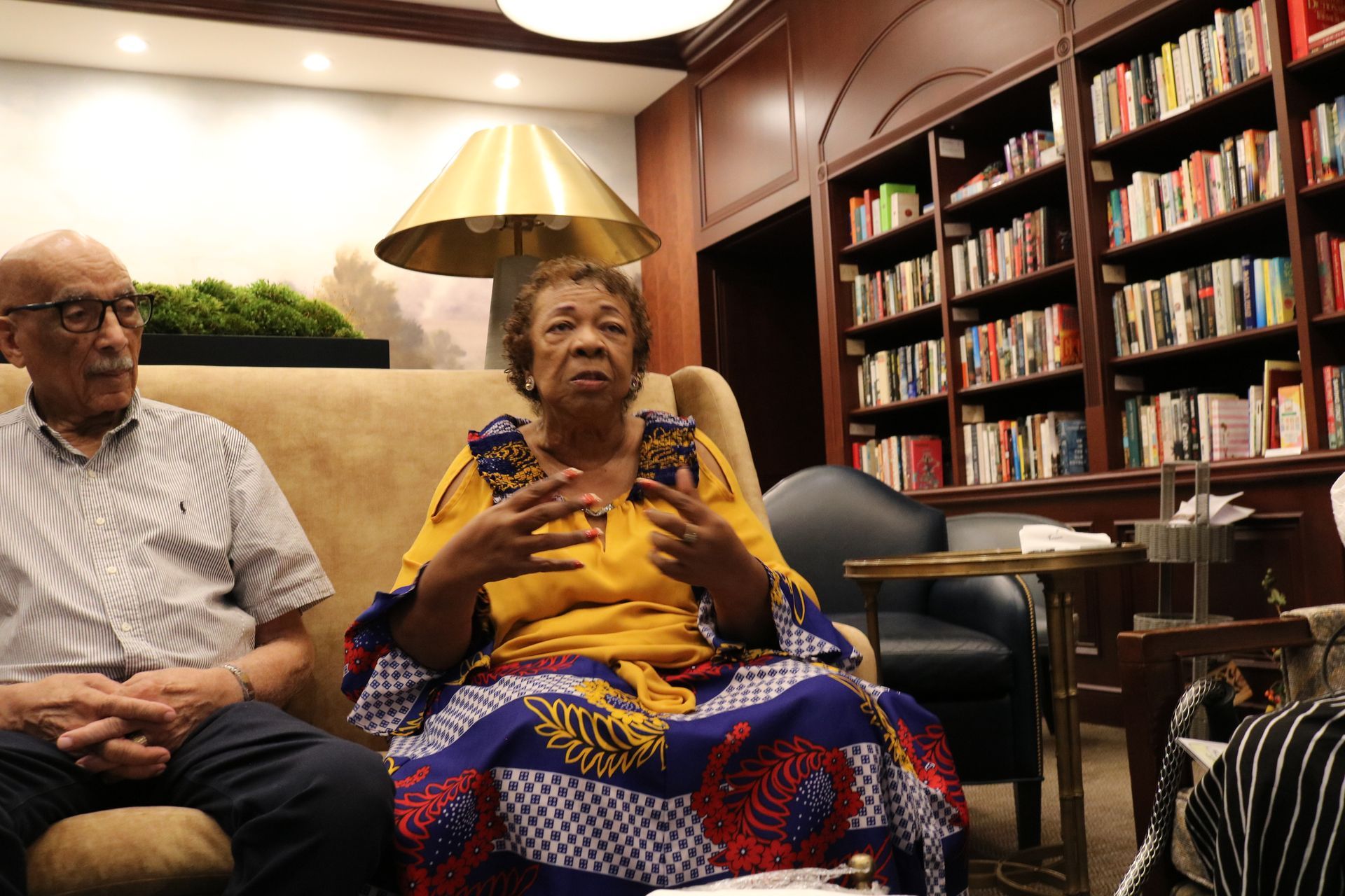 Woman in yellow and patterned dress gesturing, seated, beside a man; books, lamp, and shelves in background.