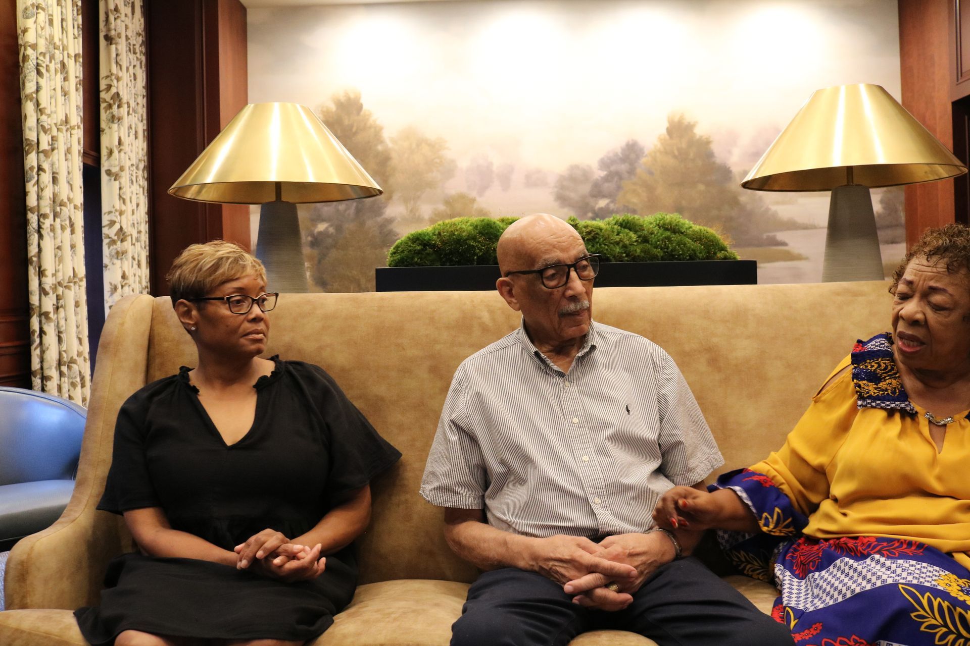 Three people seated on a brown sofa, conversing. Background features landscape painting and gold lamps.