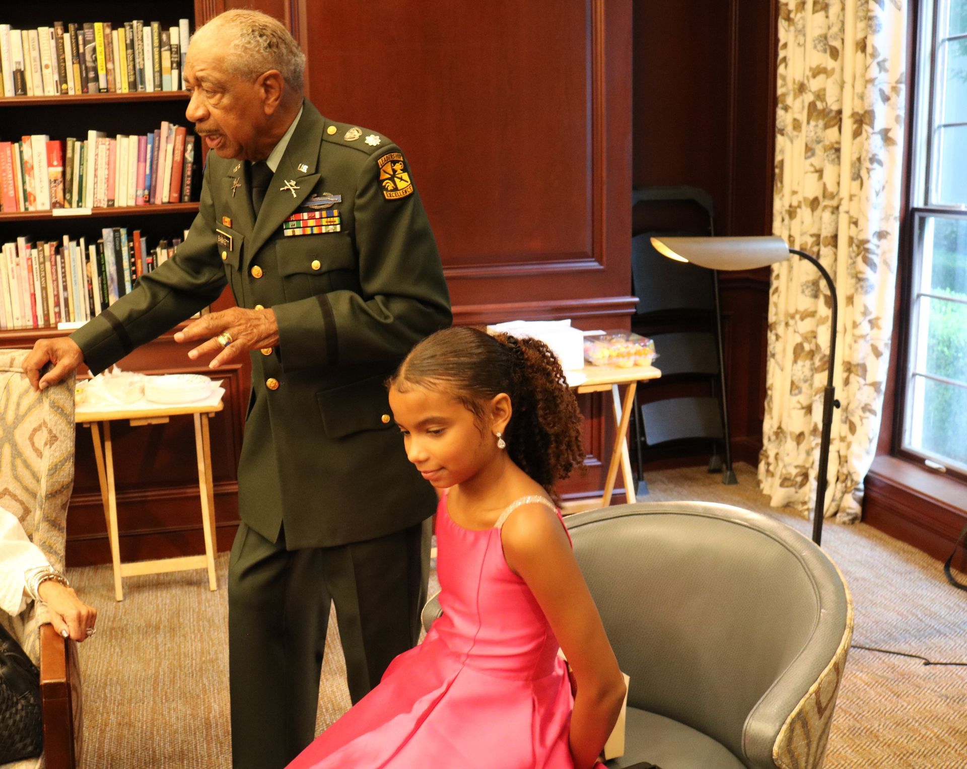 Man in military uniform speaks to girl in pink dress; bookshelf background.