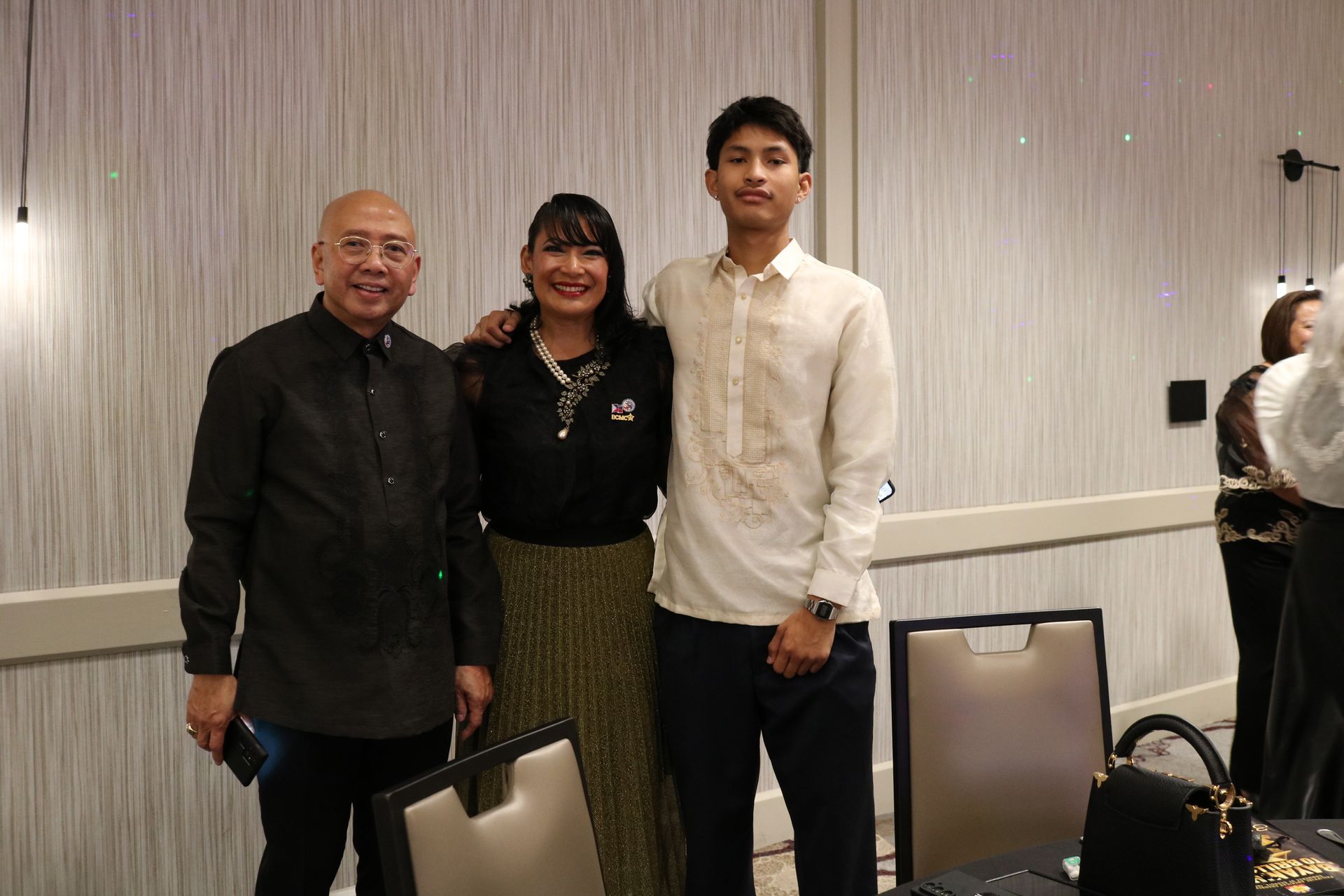 Three people posing for a photo indoors; an older man, a woman with her arm around the man, and a young man in formal attire.