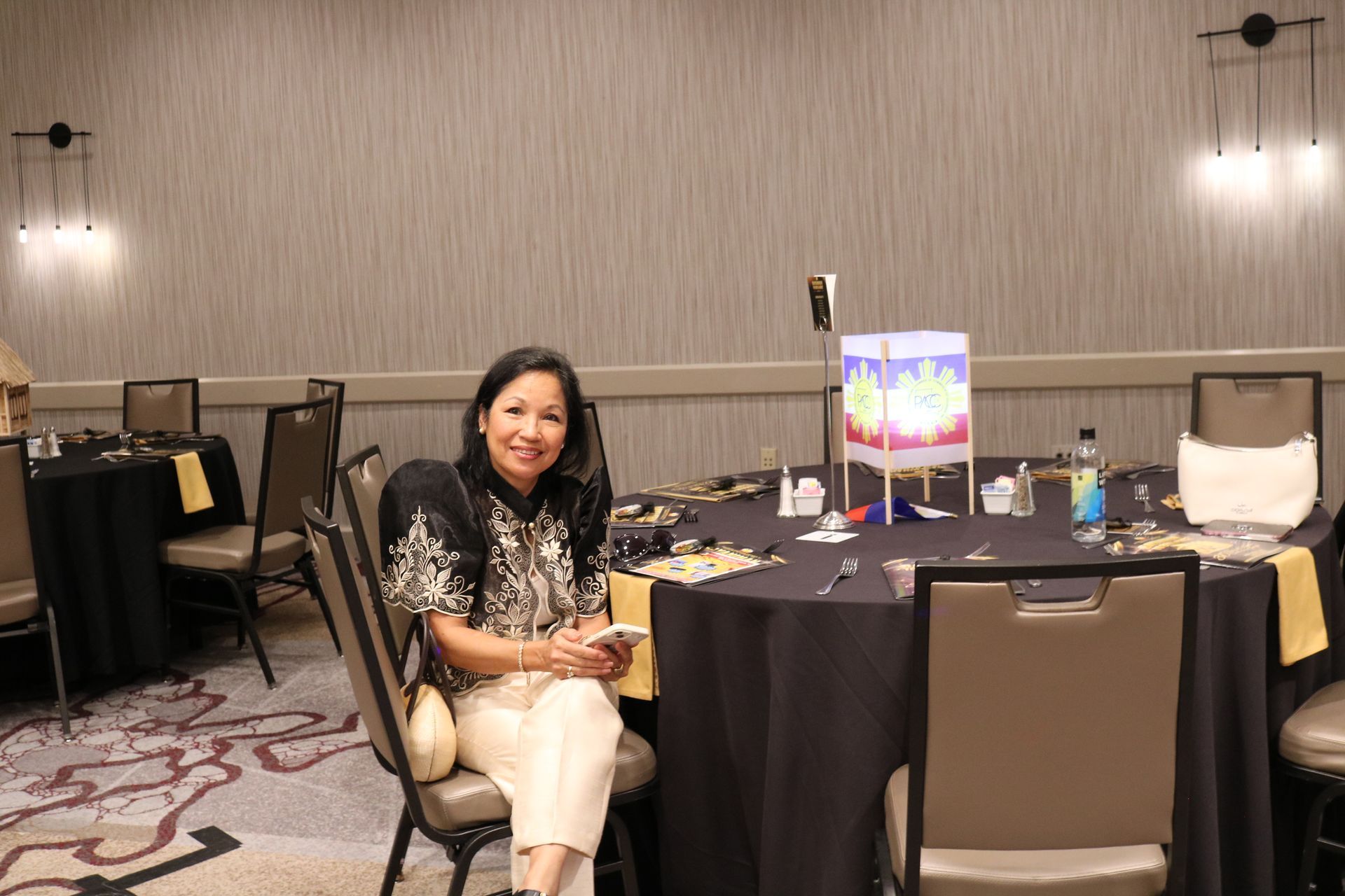 Woman seated at a table in a banquet hall, smiling, holding phone. Table set with decorations.