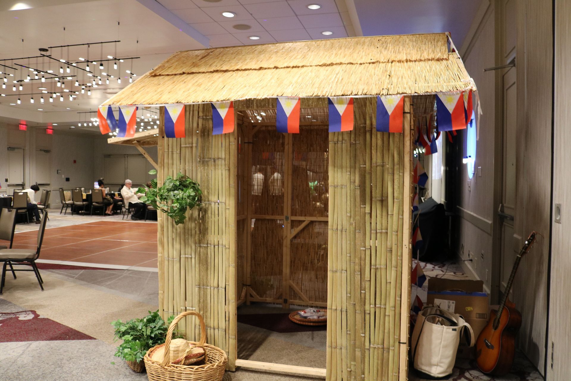 A bamboo hut decorated with Philippine flags stands indoors, with a basket and guitar nearby.