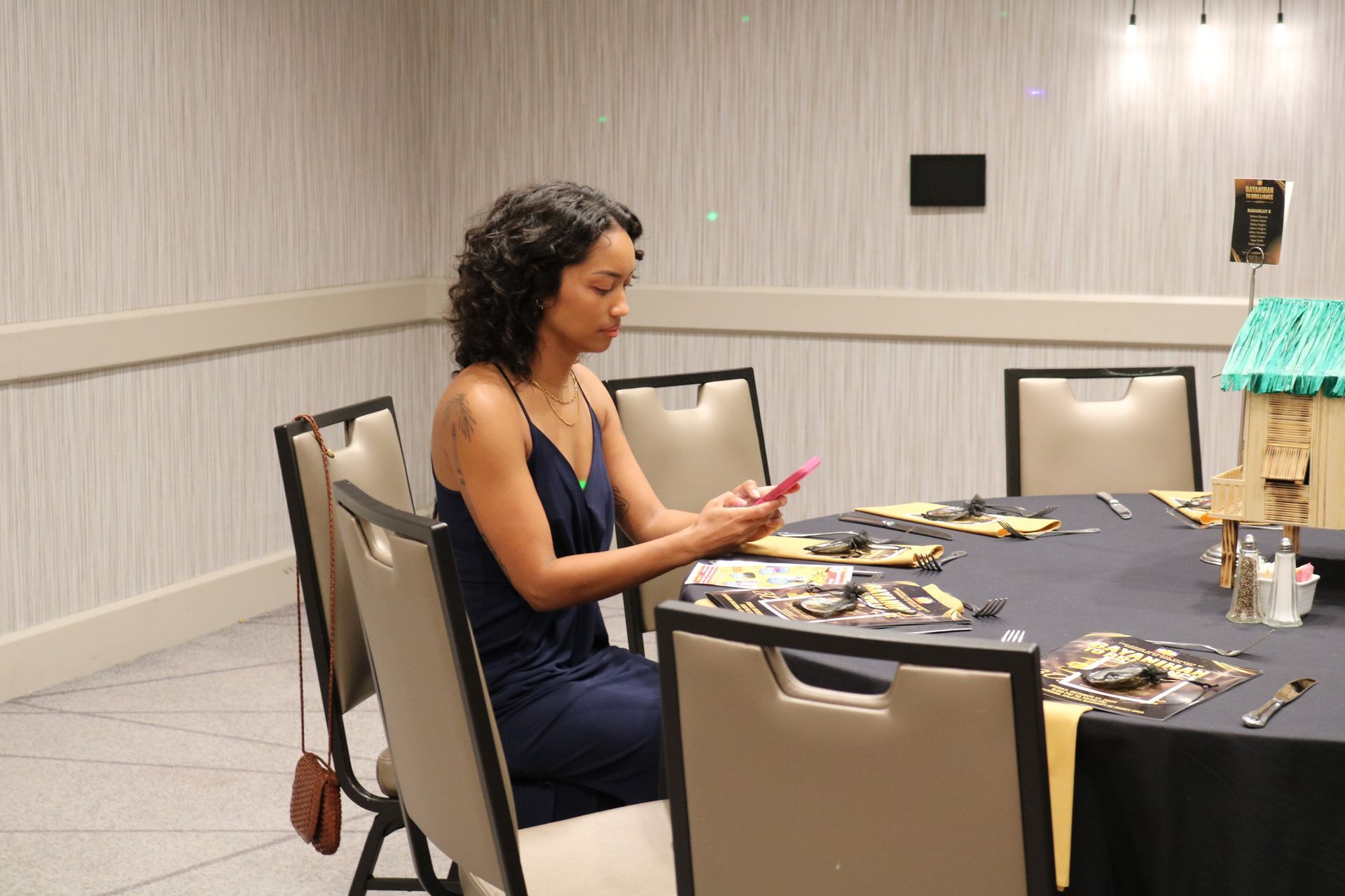 Woman in a blue dress sits at a table, looking at her phone. The table is set with decorations.