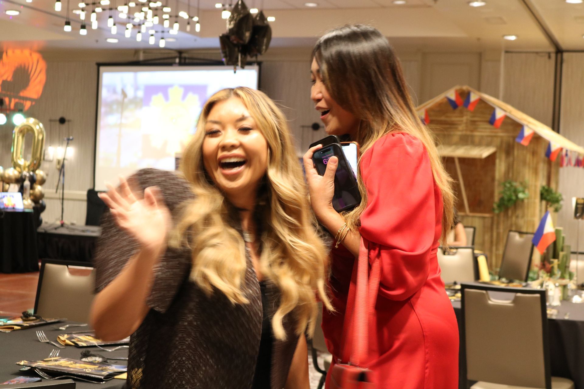 Two women at an event; one waves and smiles, the other smiles and holds a phone. Room setting.