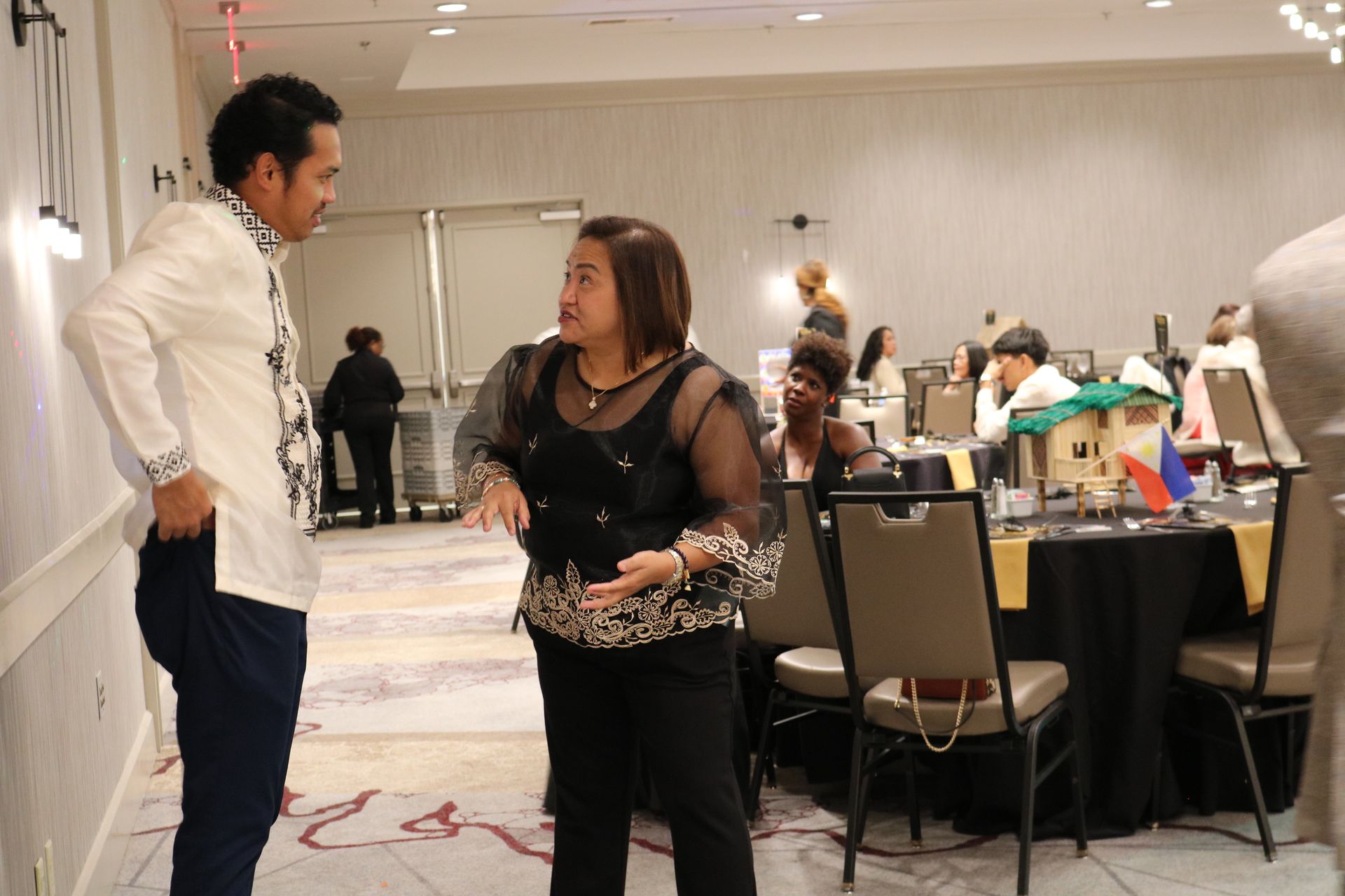 Two people conversing in a brightly lit room with tables and other people in the background.