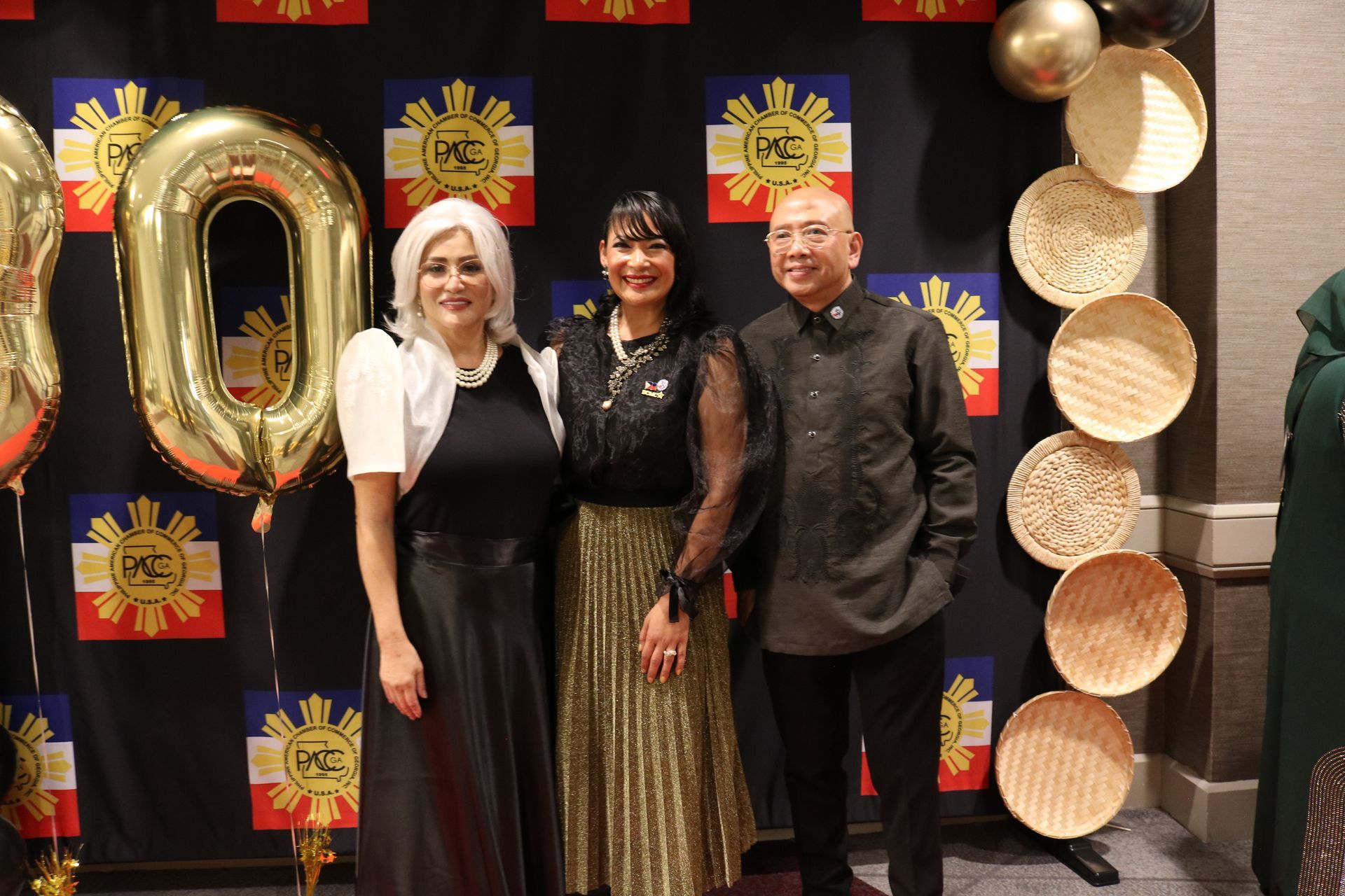 Three people pose in front of a backdrop with balloons and flags. Two women and a man are smiling.