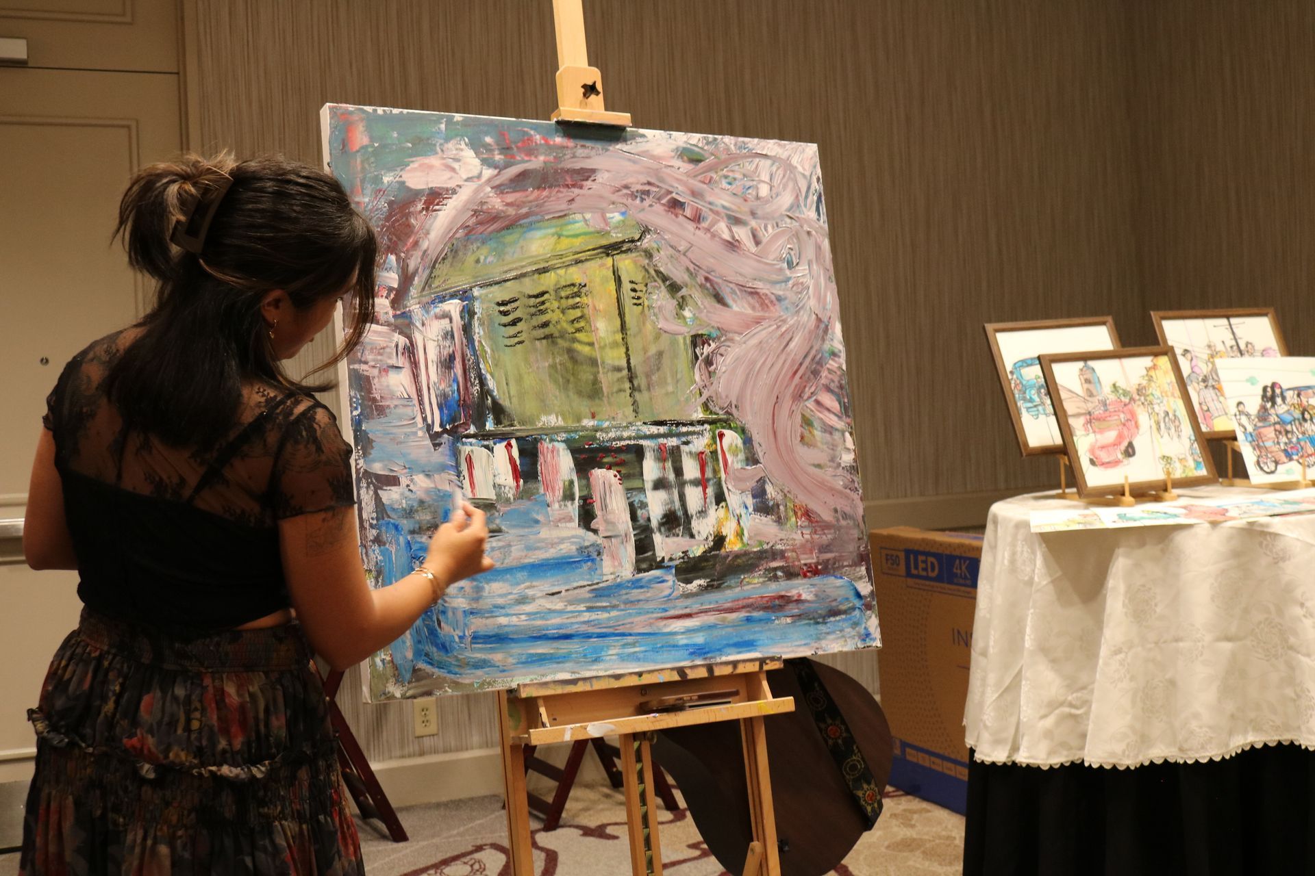 Woman painting at an easel. Colorful abstract painting in progress. Other framed art on a nearby table.