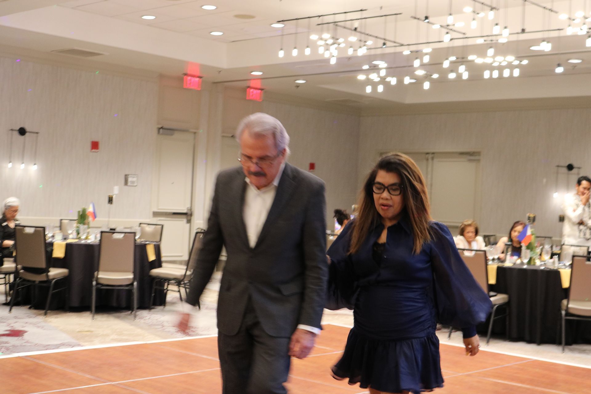 Two people walking on a dance floor in a ballroom. Man in a suit, woman in a blue dress. Tables with guests visible in background.