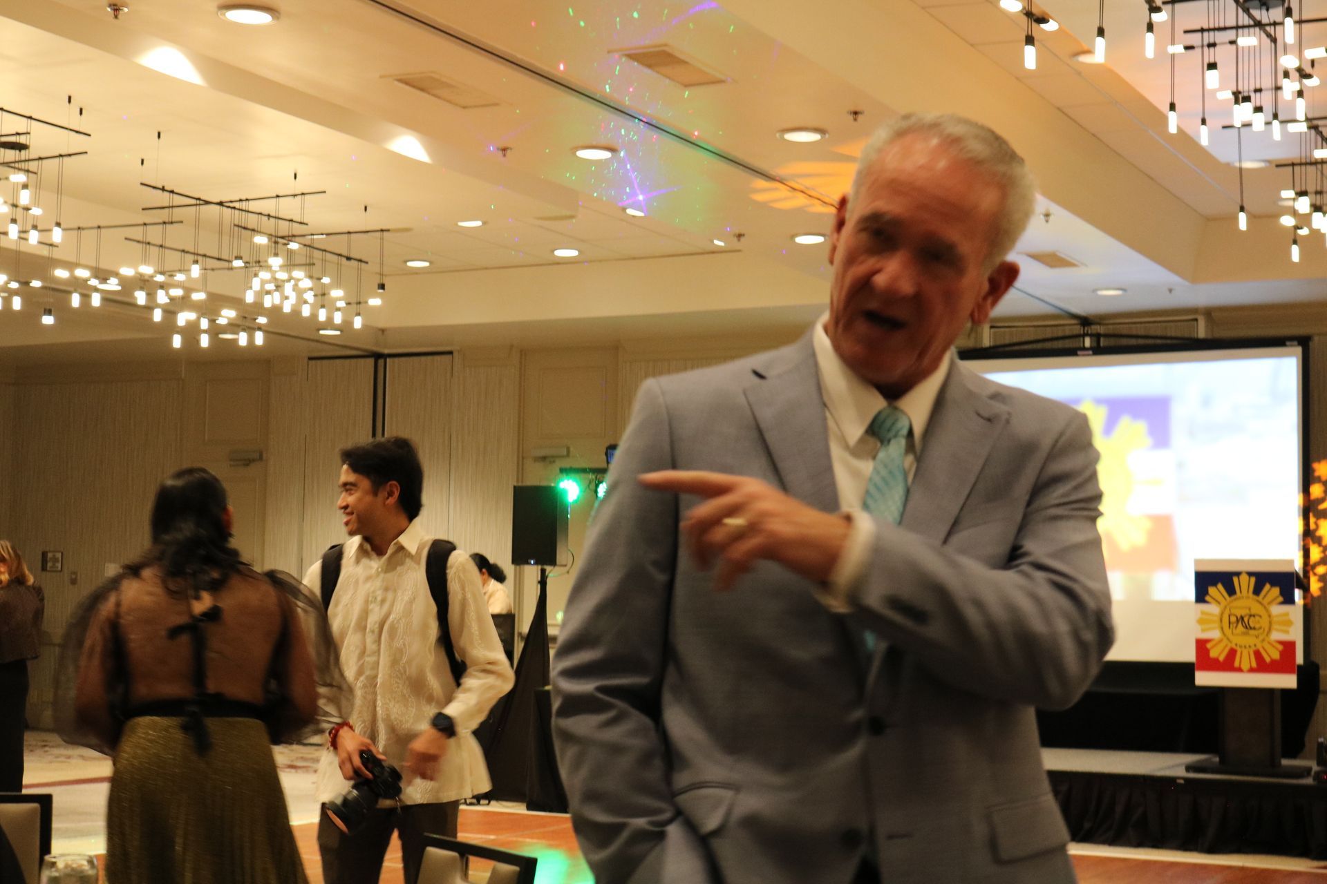 Man in suit points, speaking at event. Attendees and projection screen in background.