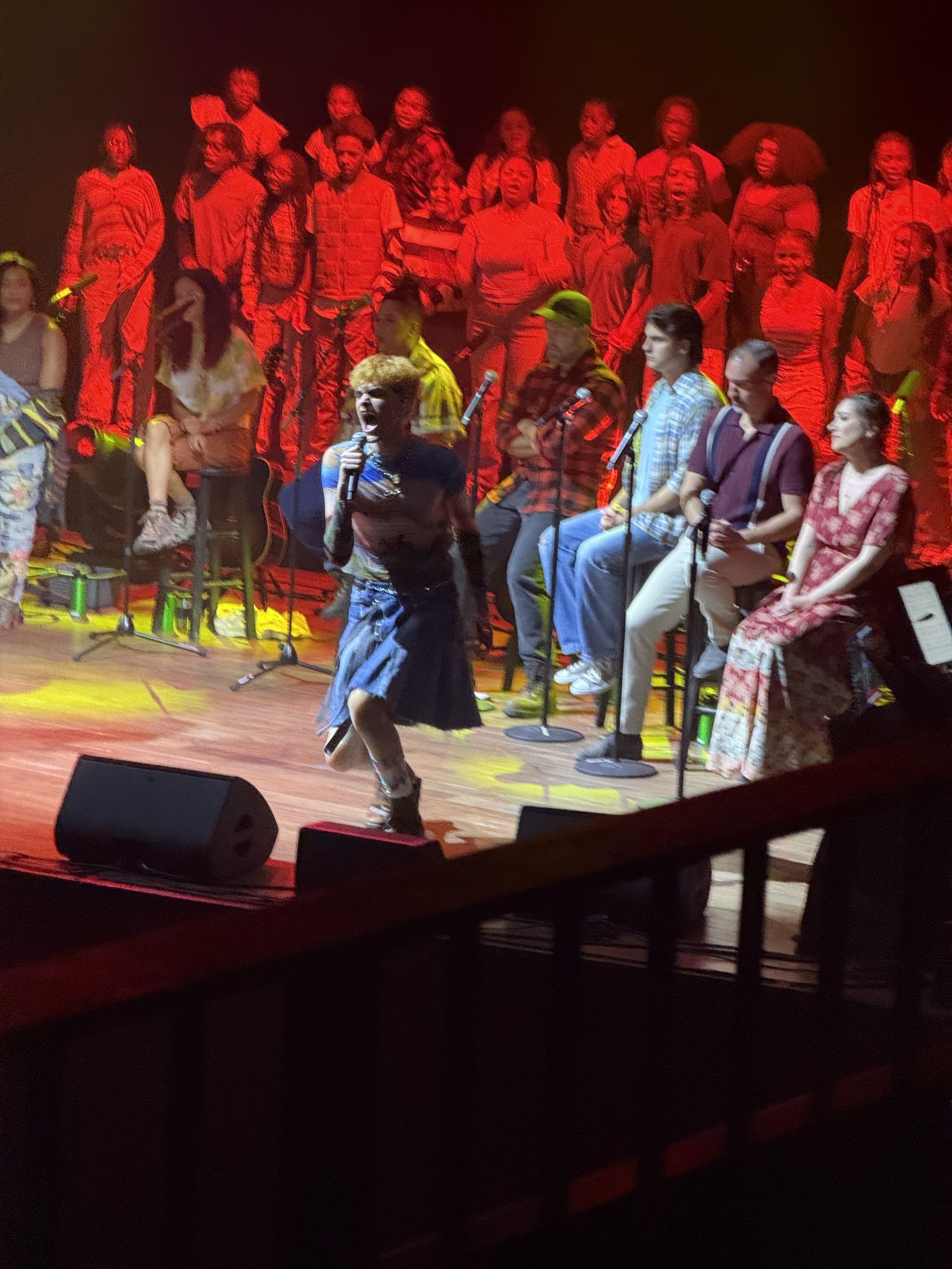 A person singing on stage with a large choir behind them under red lighting.