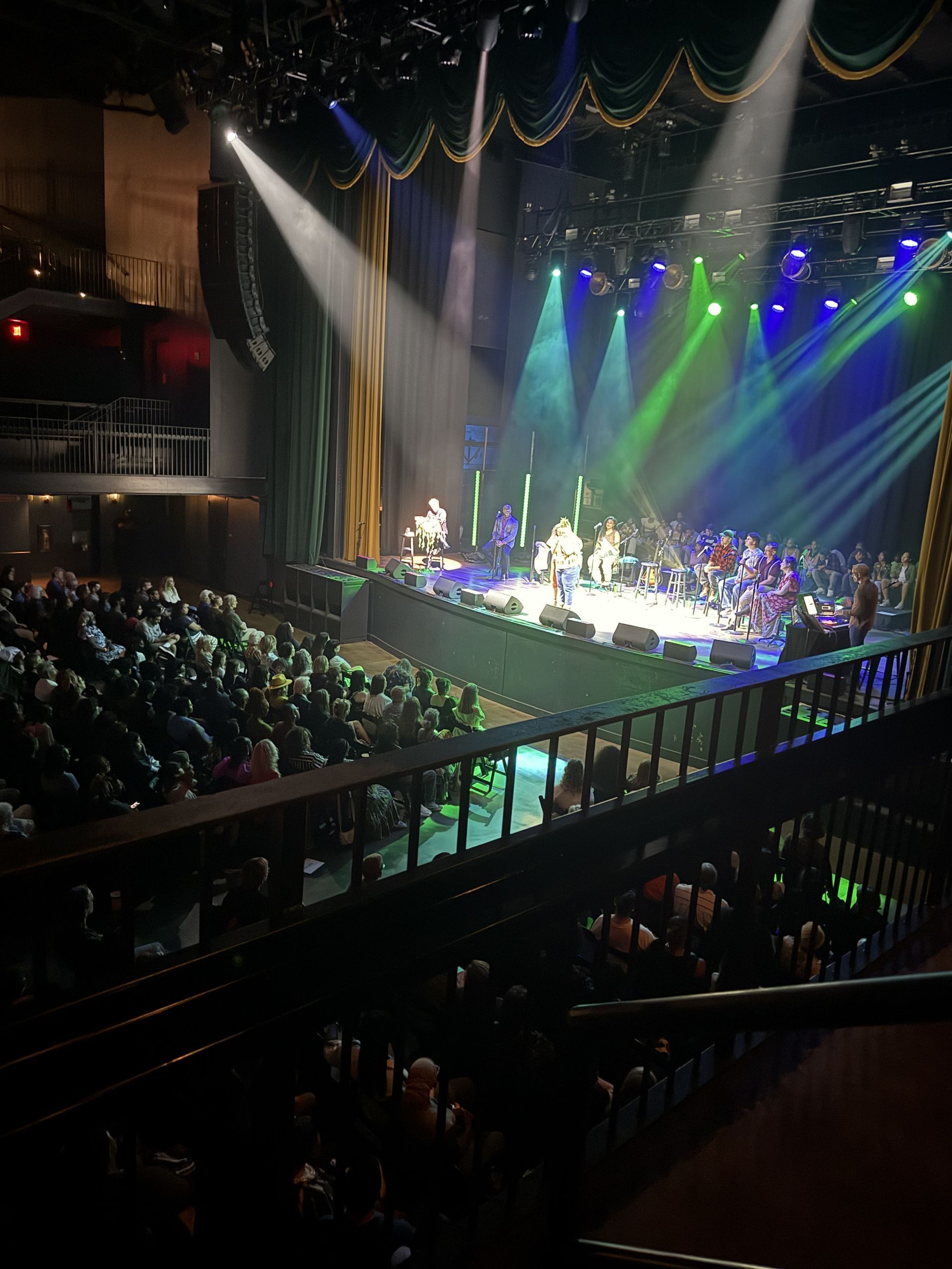 Stage with band performing, illuminated by colorful spotlights. Audience in a dark theater watches.
